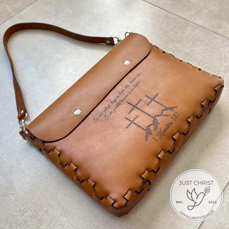 A tan leather Bible cover with a short strap seems to be on grey tiles. Three crosses with a verse are engraved.  The cover is closed with two magnetic snaps. The cover boasts a lace effect.