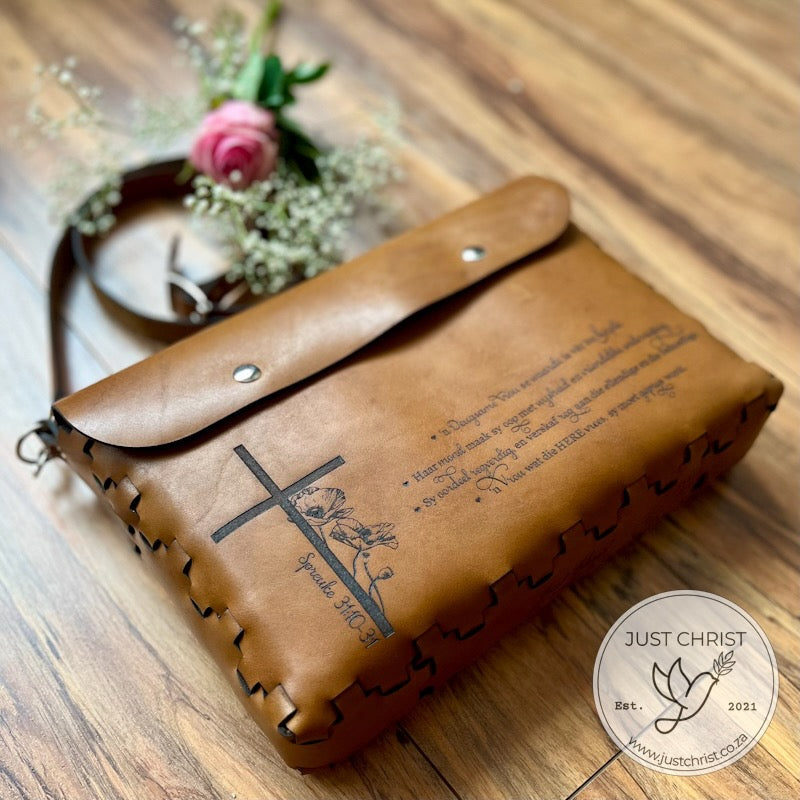 A tan leather Bible cover with wood veneer in the background.  A pink rose lays on the strap the cover. A cross with a flower is engraved, along with additional words. The cover is closed with two magnetic snaps.