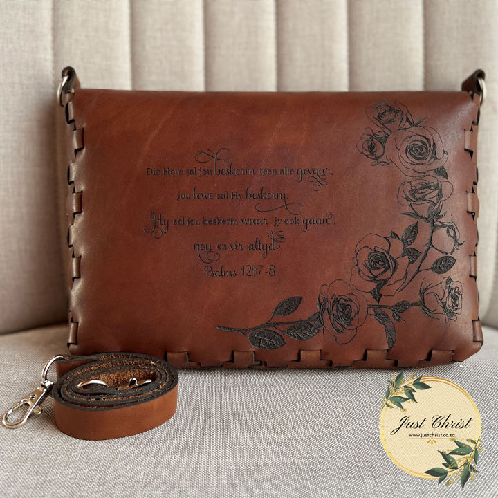 The backside of a red brown bible cover is shown,  along with the accompanying strap. Roses and a verse is engraved. 