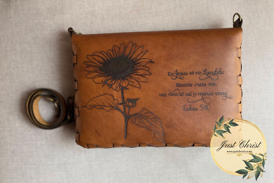 A bible cover with a strap, along with a large engraving of a sunflower and biblical verse.