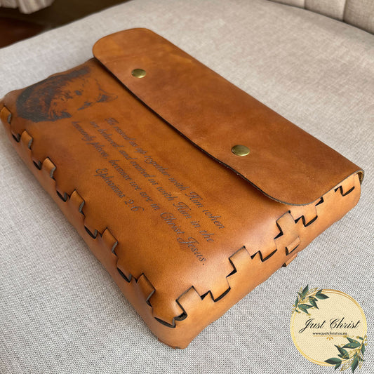 A bible cover with a no-stitch, lace effect is placed on a material background. There is a flap on the front, below the flap is an engraving of a bear and a biblical verse. 