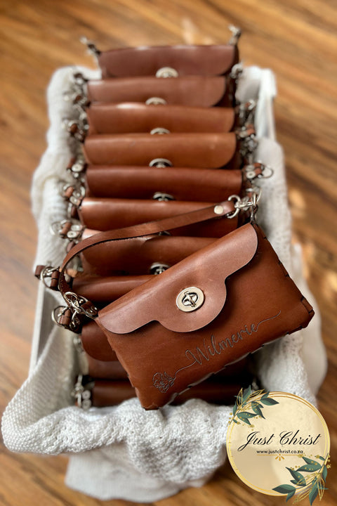 Several leather clutches is placed in a basket, with one leather clutch lifted higher than the others. There is a name engraved on the front with a flower and a stem and a short strap is also evident.