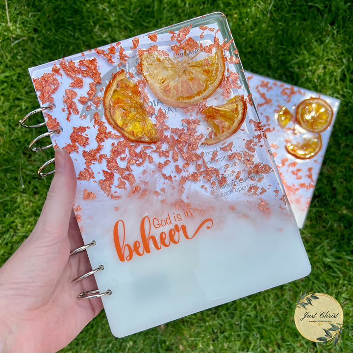 A resin notebook cover that has copper flakes in the top right corner and dried orange, the bottom left ihas white in the resin and "God is in beheer" is in orange vinyl. The notebook is held. There is another notebook partially in the background on the grass. 