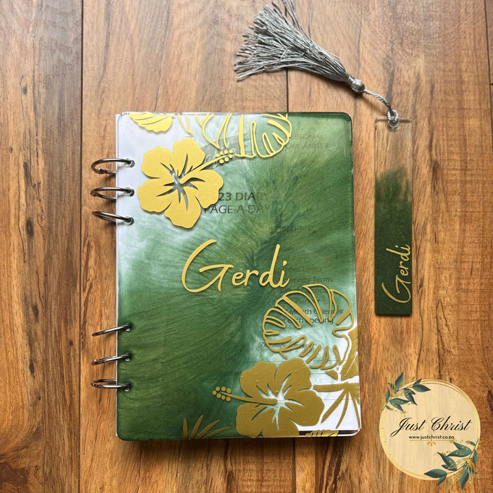 A notebook with a resin cover along with a bookmark is placed on wood laminated flooring. There is green in the centre of the notebook, moving outwards to the sides of the notebook. There are also gold flowers on the cover with a personalized name. 