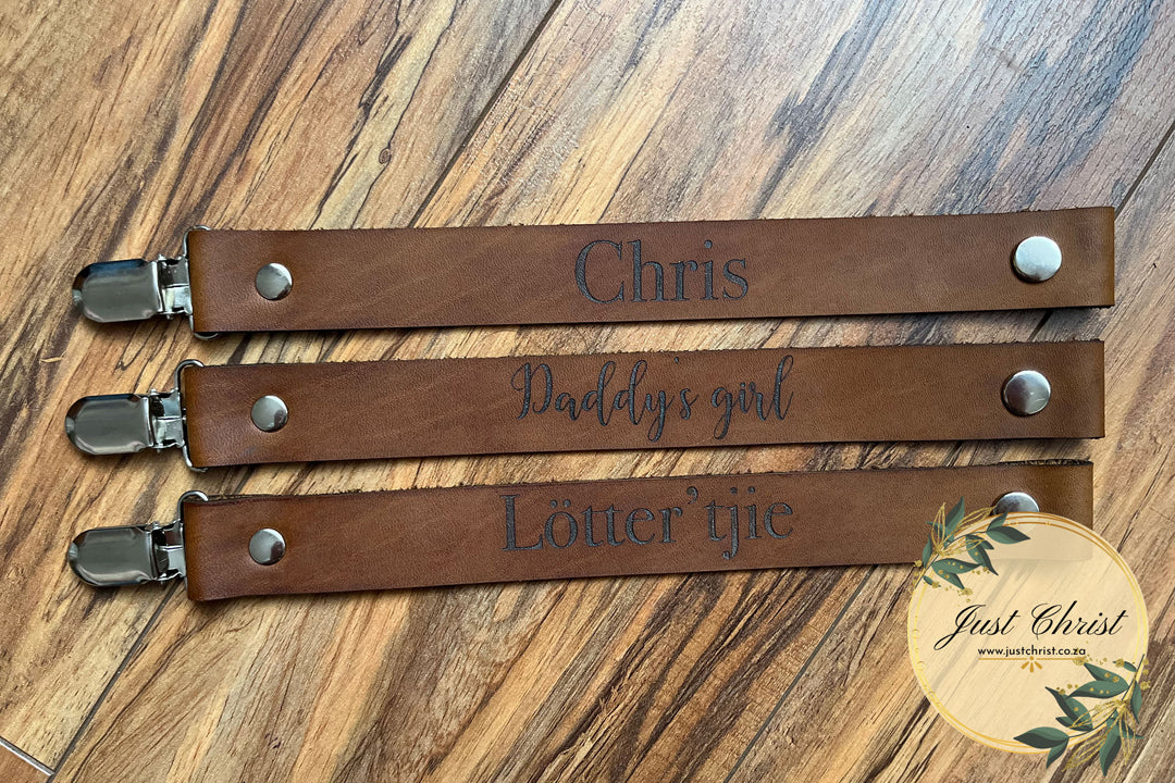 Three leather dummy clips are placed on laminated wood flooring. The Top dummy clip has "Chris" engraved, the middle dummy clip has "Daddy's girl" engraved and the bottom dummy clip has "Lotter'tjie" engraved.