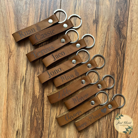 Ten leather loop keyrings are placed on a wooden background. The rings are 30mm and has a silver finish. Each leather keyring is engraved with a different name.