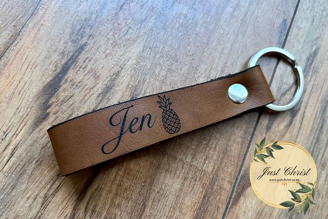 A leather loop keyring is placed on a wooden background. It has a silver keyring and "Jen" with a pineapple is engraved.