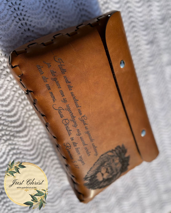 A beige leather bible cover taken from the front. The cover is on a white blanket. A verse and lion (cat) is engraved on the front side of the cover. Two magnetic snaps is also shown, which is part of the cover.