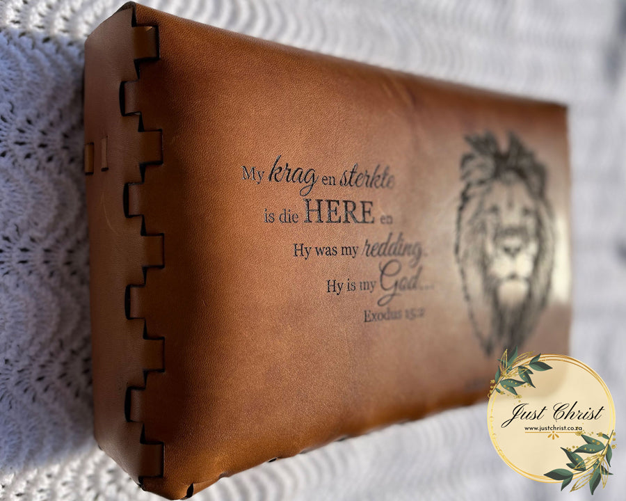 The back-side of a beige leather bible cover is shown in the image. The cover is placed on a white blanket and a verse and lion (cat) is engraved on the cover.