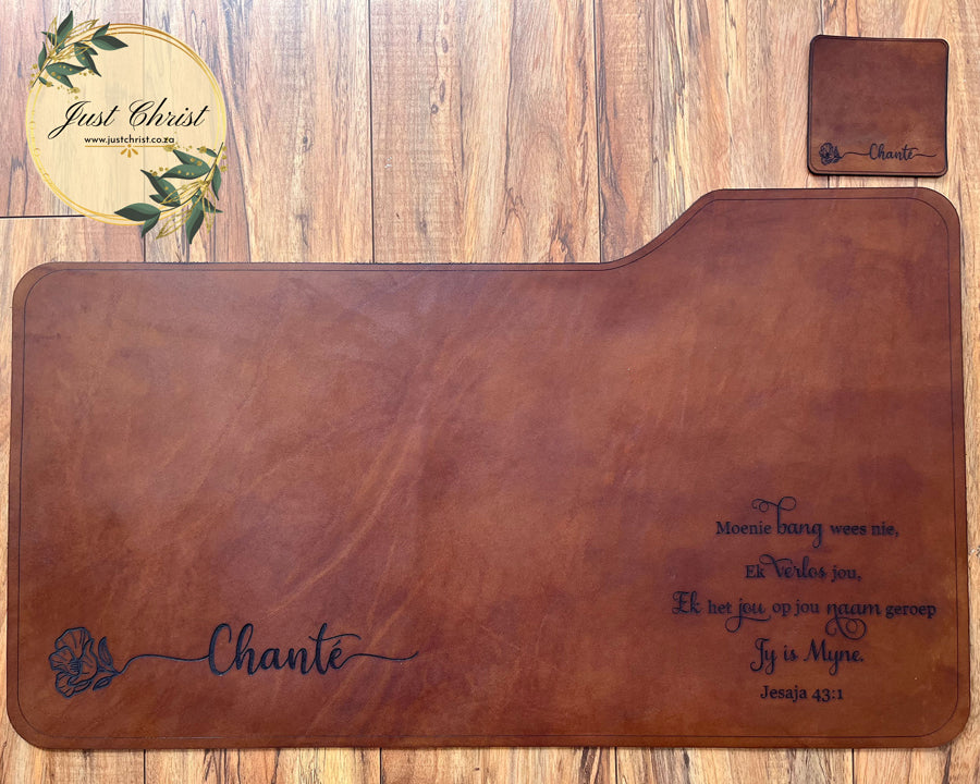 demonstrated in the image. The deskpad is on wood flooring and there is a matching coaster to the right of the deskpad. The engraving is a name on a flower stem, as well as the verse "Jesaja 43:1" in Afrikaans at the bottom right corner of the leather deskpad.