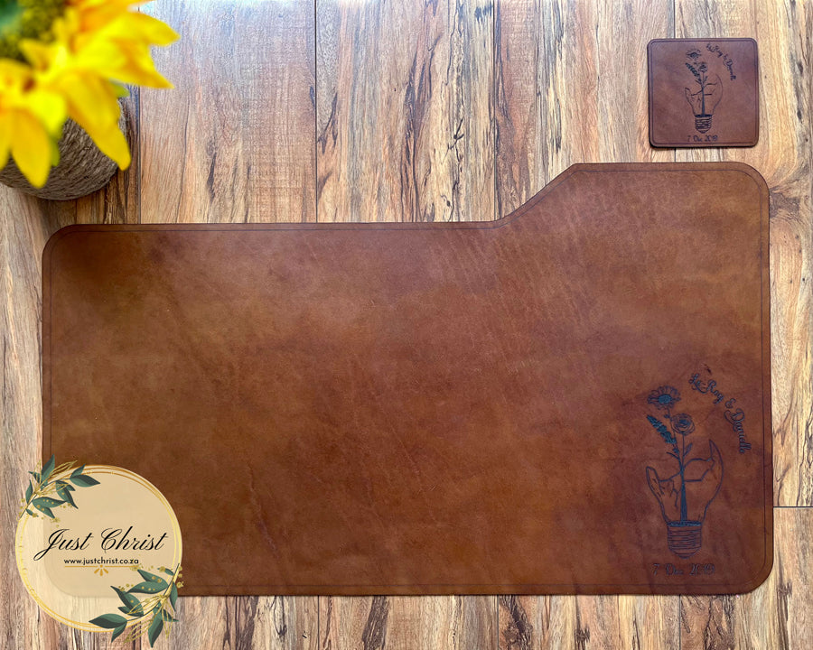 A full photo of the leather deskpad is demonstrated in the image. The deskpad is on wood flooring and there is a matching coaster to the right of the deskpad. The engraving is a broken lightbulb with wildflowers growing from the base. There is a date at the bottom of the engraving. 