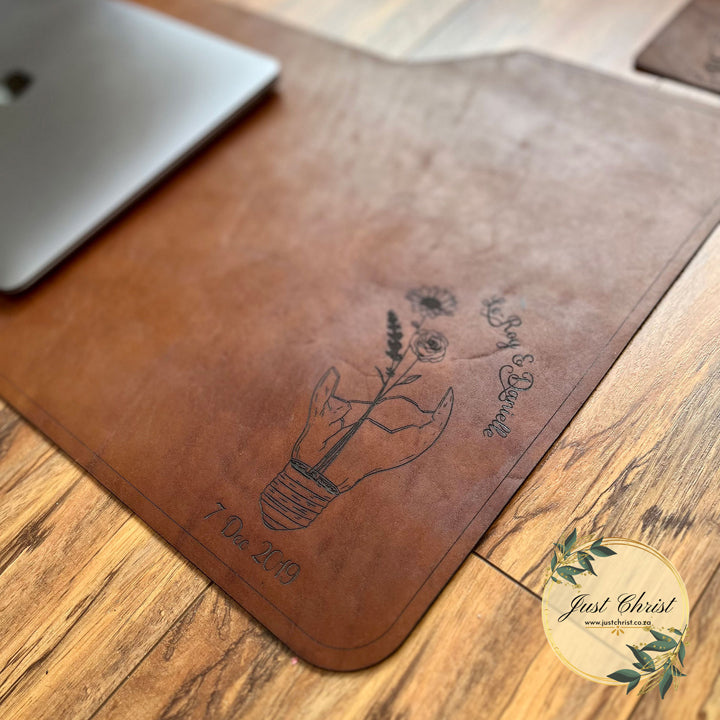 A medium deskpad was placed on laminated wood flooring. There is a laptop on the deskpad. There is an engraving of a broken lightbulb engraved on the leather. The image is taken from an angle. 