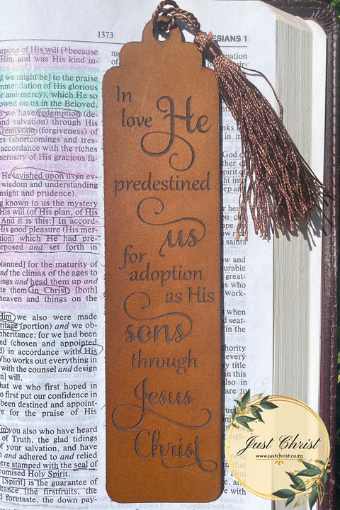 A beige leather bookmark with a brown tassel is strategically placed on an open bible, it appears to be Ephesians 1 in the background. The leather bookmark has the following engraved: "I love He predestined us for adoption as His sons through Jesus Christ."