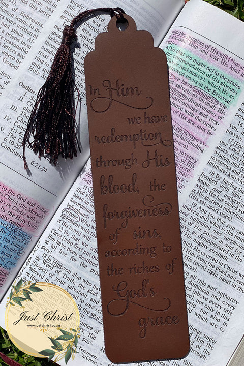 A leather bookmark is placed on a Bible page, the bookmark has an accompanying brown tassel and the following is engraved: "In Him we have redemption through His Blood, the forgiveness of sins according to the riches of God's grace."