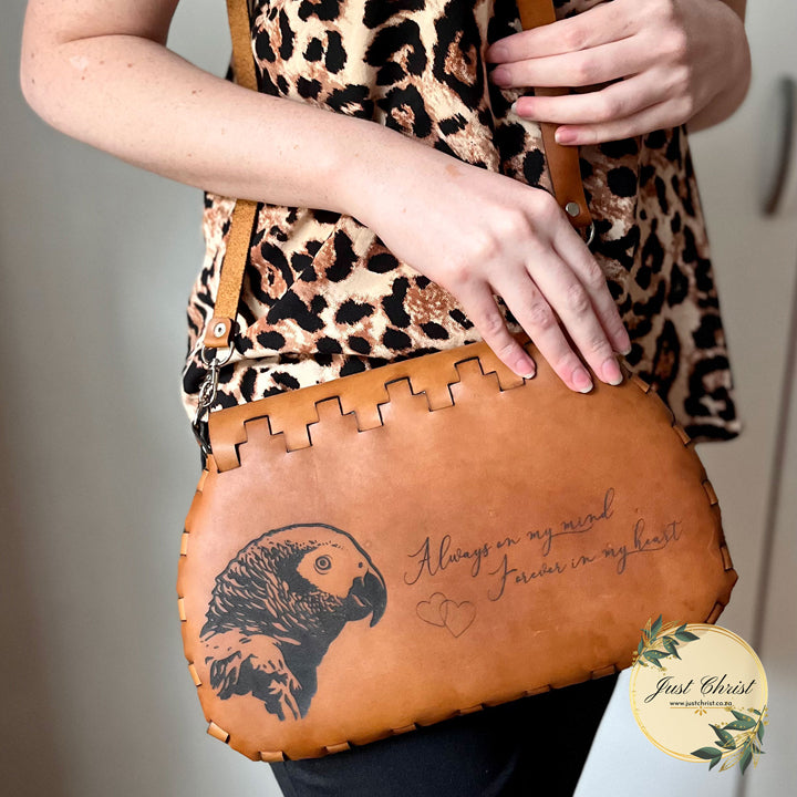 The back side of a large purse is shown. A lady has the purse over her shoulder, and she is holding on to the strap with one hand and the purse with the other hand. She has a leopard print shirt on. The purse is beige and the sides have a lace effect. An African Grey bird is engraved on the leather with the following saying: "Always on my mind, Forever in my heart."