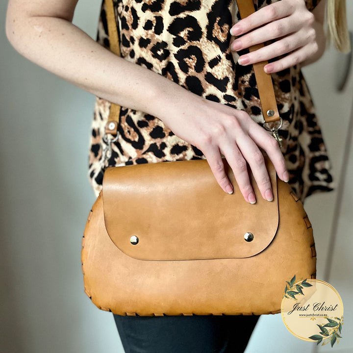 The front side of a large purse is shown. A lady has the purse over her shoulder, and she is holding on to the strap with one hand and the purse with the other hand. She has a leopard print shirt on. The purse is beige and the sides have a lace effect.