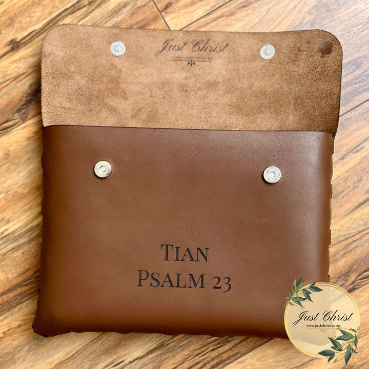 A leather Bible cover suitable for standard bibles is placed on wood laminated flooring. The Bible cover has a flap which is opened and engraving of a name and a verse on the front. The open flap shows that the Bible cover does not have a lining but shows the rawness of the real leather.