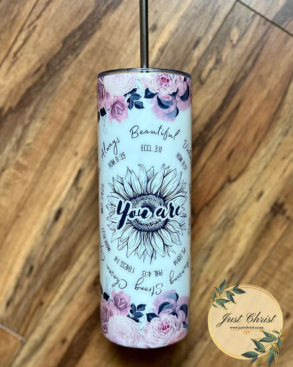 A tumbler bottle with a stainless steel straw in it is on a wooden floor. The tumbler is decorated with a wrap that lists verses and a keyword from that verse around a sunflower in a circle.